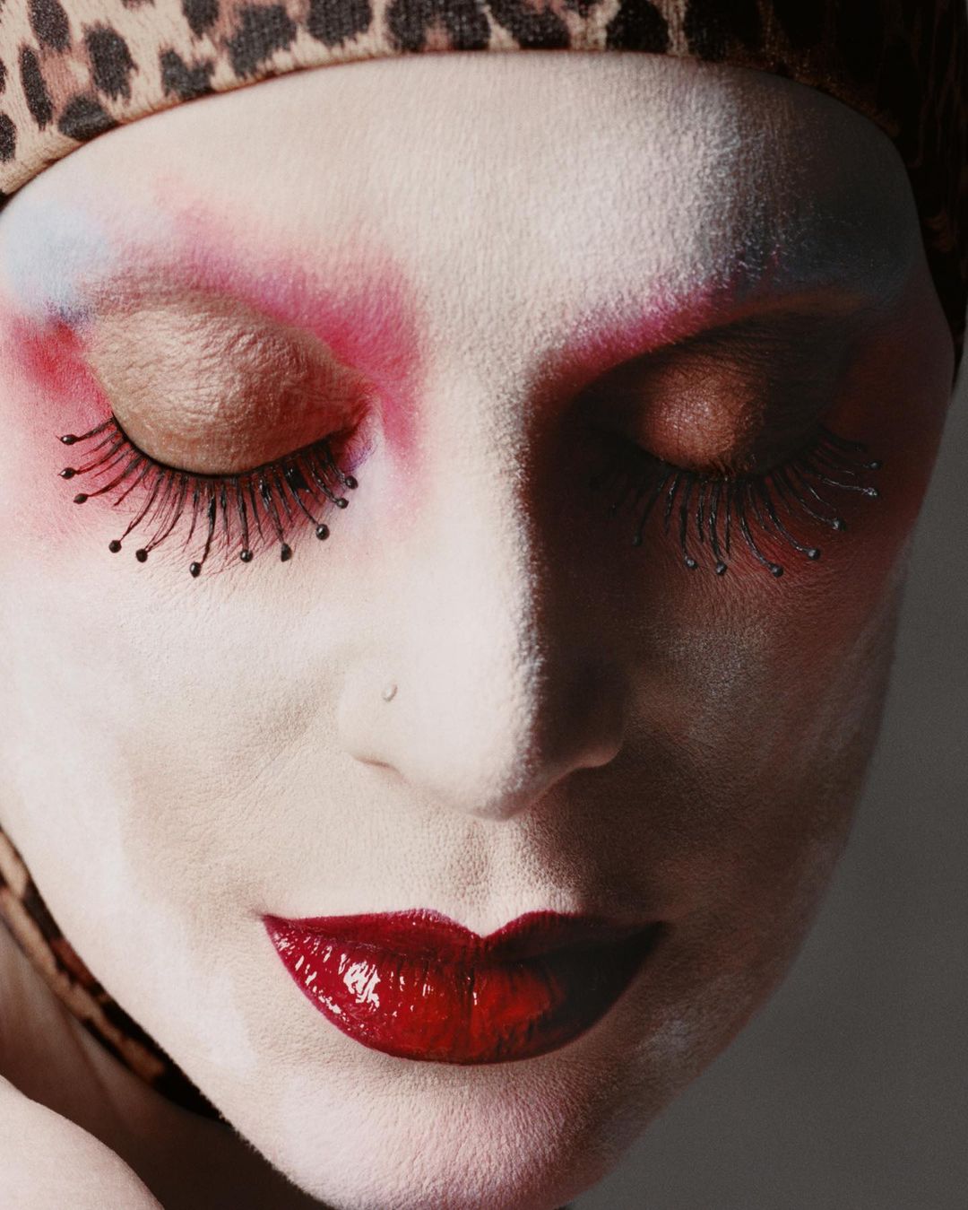Close-up portrait of a person with heavy makeup and a leopard print headscarf, eyes closed, showcasing a striking and artistic beauty look.