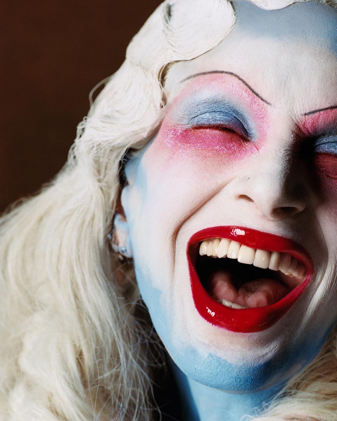 Close-up of a woman with dramatic blue and red face paint, exaggerated makeup, and a wide-open mouth in a theatrical expression.