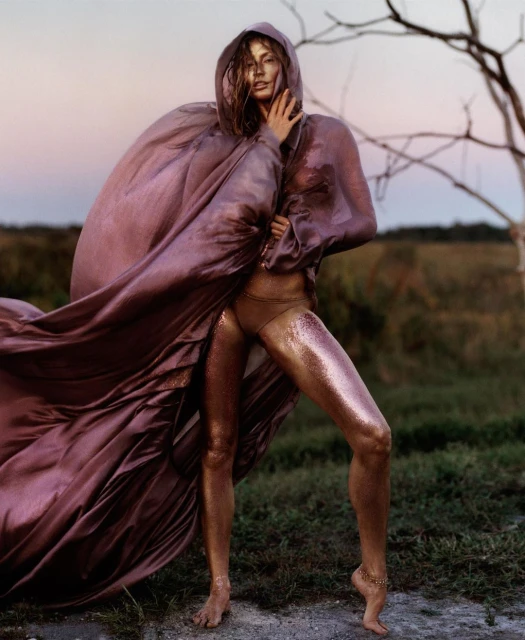 Woman in bronze body paint poses in a windswept purple hooded cloak against a field and sky at dusk in a fashion editorial shot.