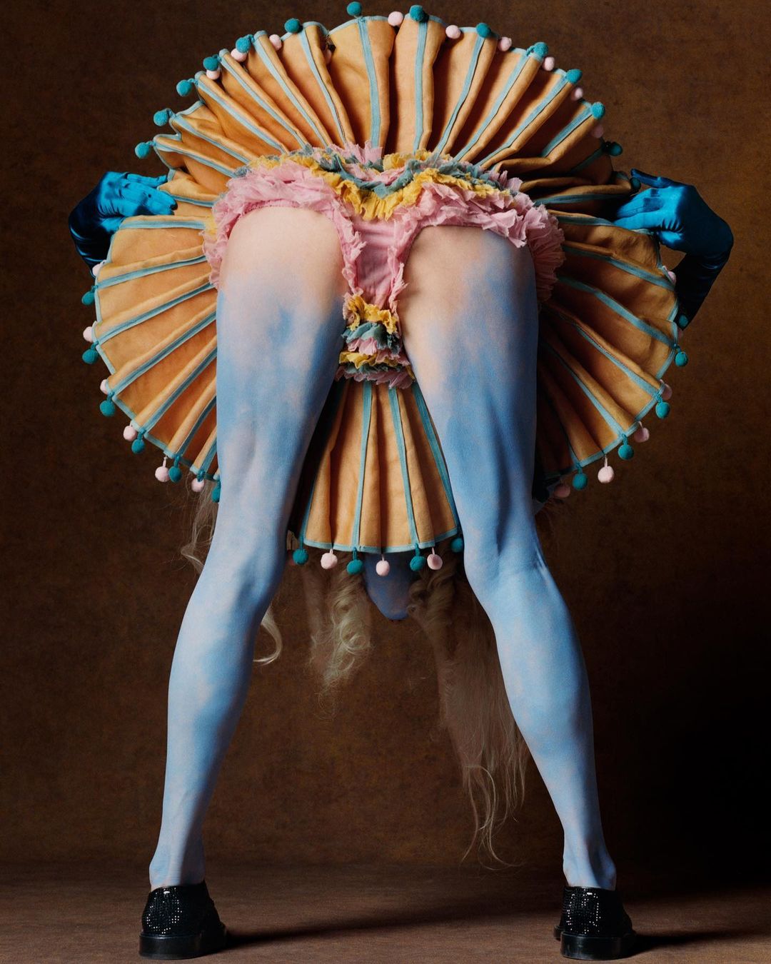 A model in an avant-garde costume bends forward, displaying their painted blue legs, ruffled skirt, and a tan ruffled collar.