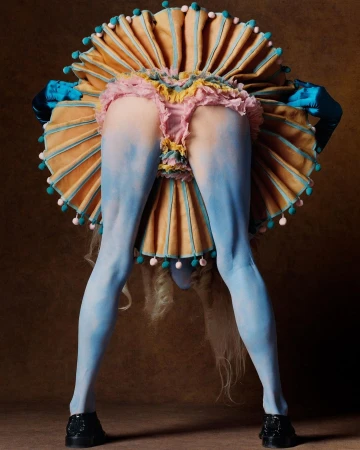 A model in an avant-garde costume bends forward, displaying their painted blue legs, ruffled skirt, and a tan ruffled collar.