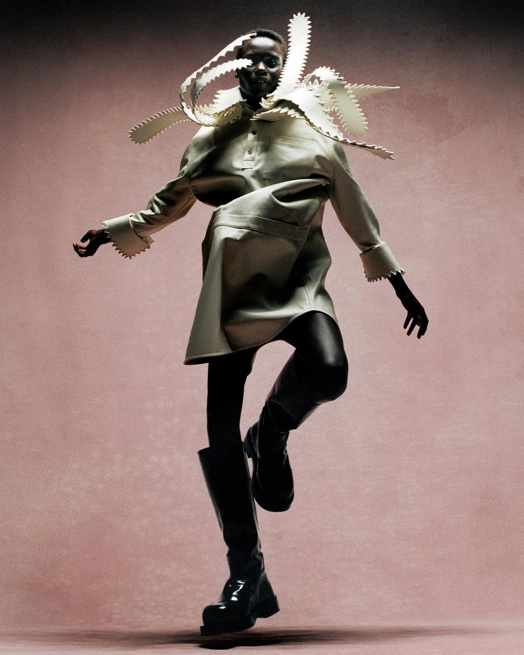 Fashion model in an avant-garde cream-colored dress and black boots poses dynamically against a textured pink background in a studio setting.