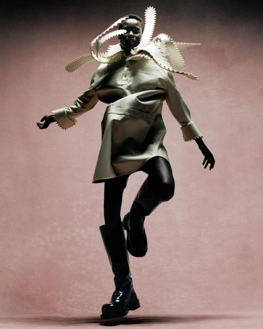 Fashion model in an avant-garde cream-colored dress and black boots poses dynamically against a textured pink background in a studio setting.