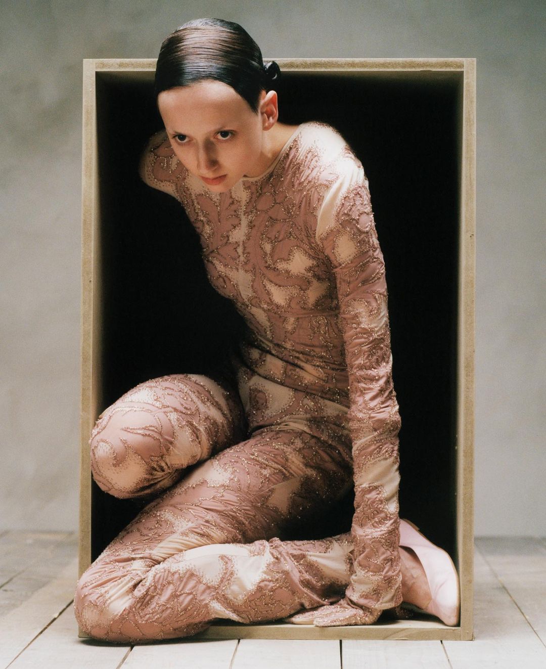 Woman in patterned body suit poses inside of a wooden box in a photo studio with a neutral background and a wooden floor.