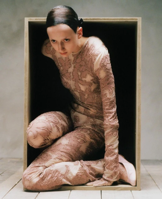 Woman in patterned body suit poses inside of a wooden box in a photo studio with a neutral background and a wooden floor.