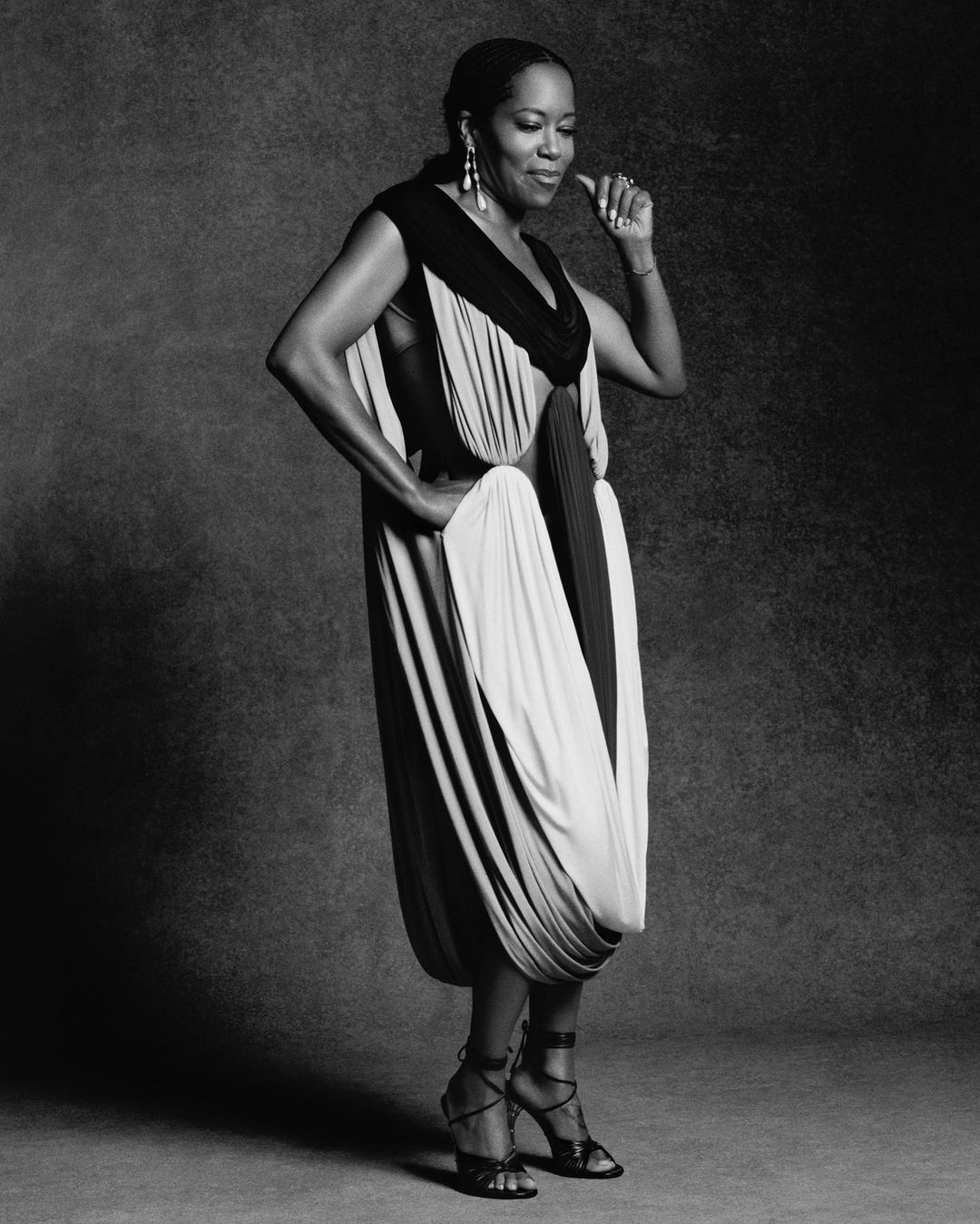 Monochrome portrait of Regina King in a draped dress and strappy heels, standing against a mottled background, showcasing elegance and style.