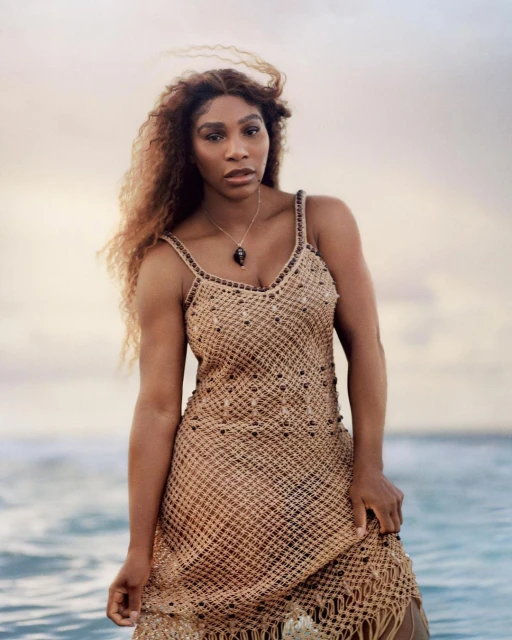 Serena Williams poses in a macrame dress on a beach with a neutral sky in the background, showcasing fashion and natural beauty.