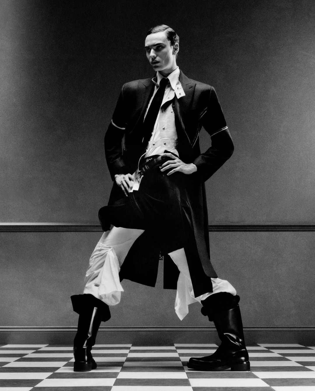 Black and white shot of a man in a coat and boots standing in a wide stance on a checkerboard floor in front of a grey wall.