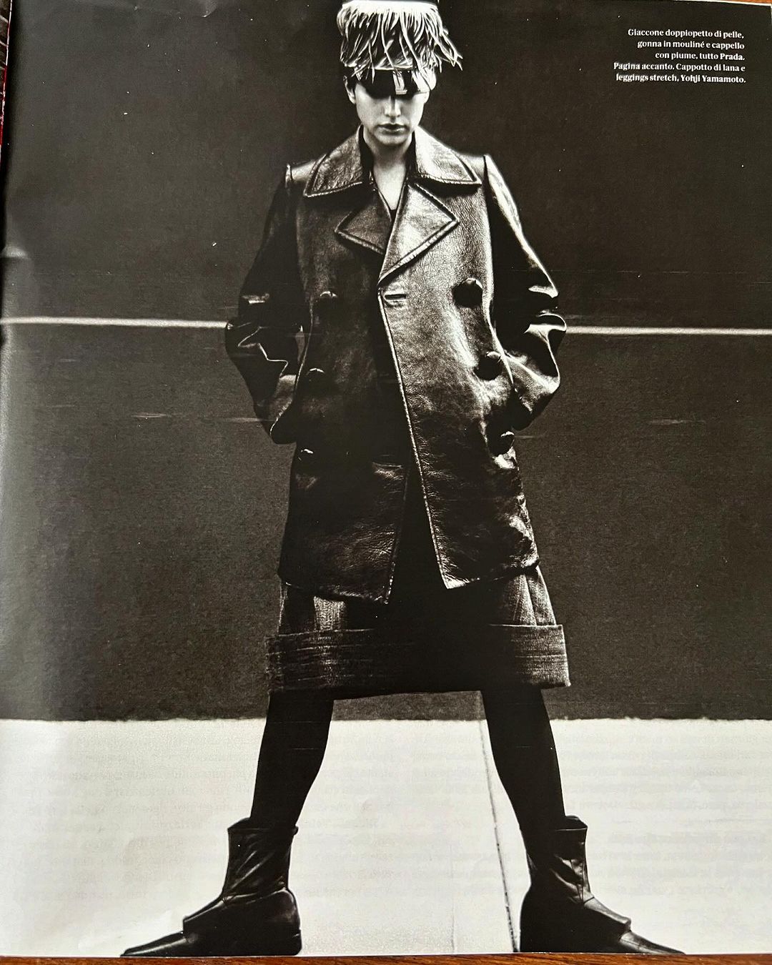 Monochrome portrait of an adult woman wearing a dark trench coat, leggings, boots, and an eye-covering hat, posing in front of a dark background.