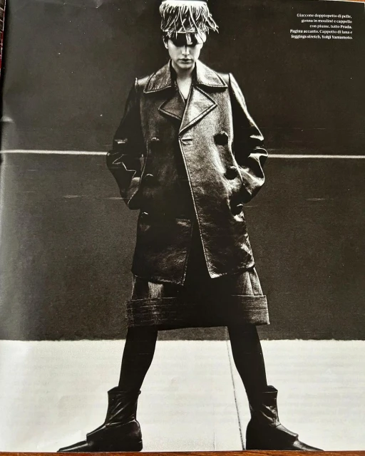 Monochrome portrait of an adult woman wearing a dark trench coat, leggings, boots, and an eye-covering hat, posing in front of a dark background.