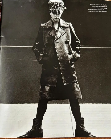 Monochrome portrait of an adult woman wearing a dark trench coat, leggings, boots, and an eye-covering hat, posing in front of a dark background.
