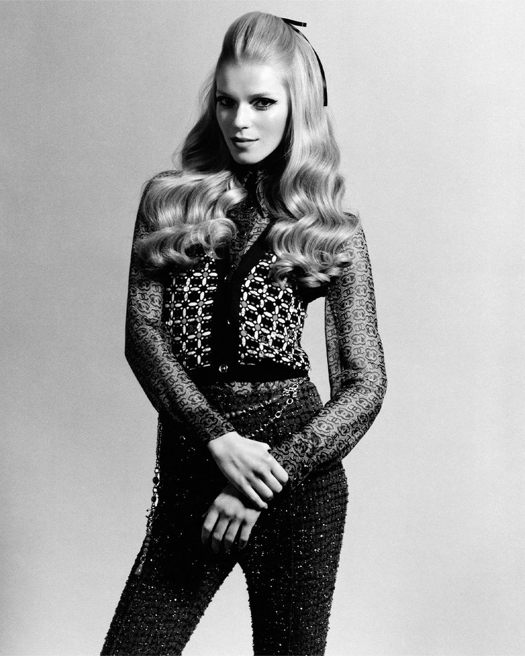 A woman with blonde hair and a black bow poses in a black and white photo. She wears a patterned top, sheer sleeves, and matching pants.