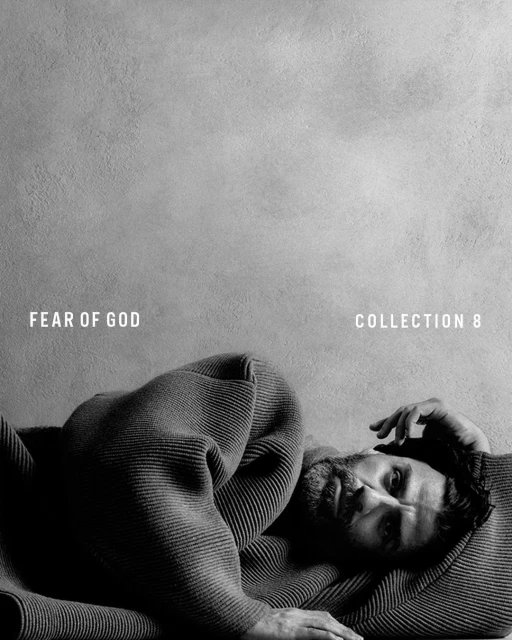 Monochrome portrait of a bearded man lying down wrapped in a knit sweater, eyes open, in front of a textured grey backdrop with brand text.
