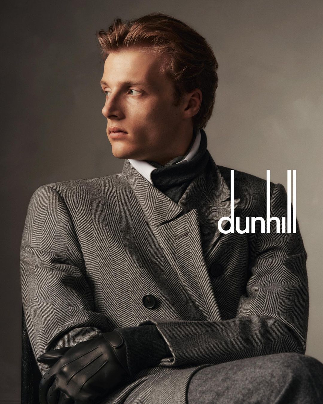 Portrait of a fair-skinned adult male model in a grey overcoat and black leather gloves, looking off to the side in soft light.