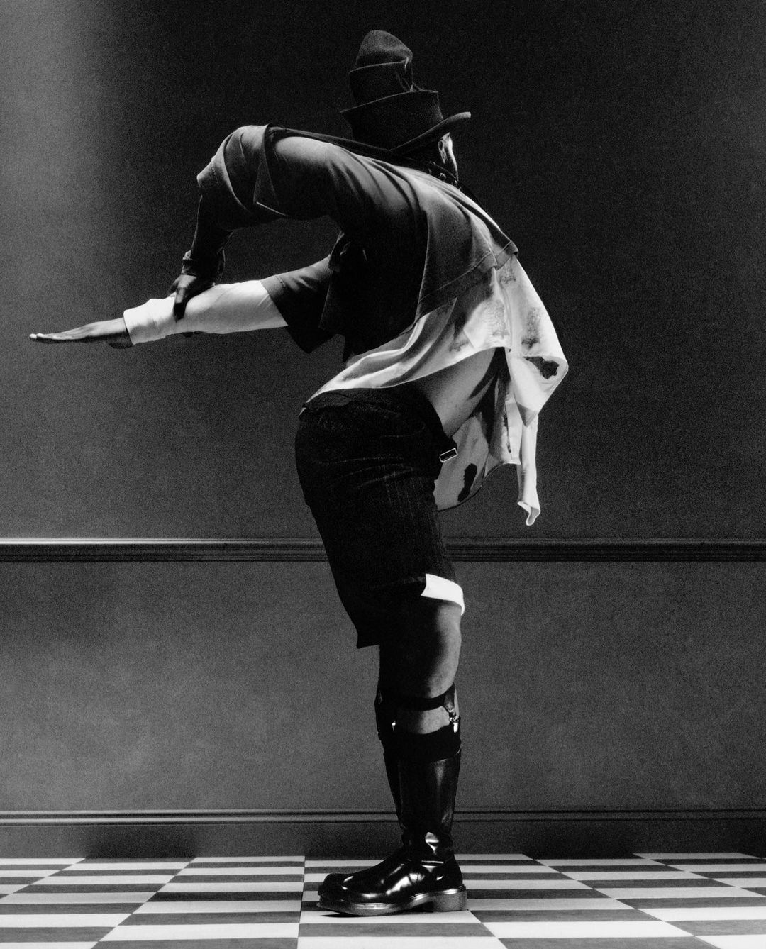 Monochrome full-length portrait of a person with a hat and boots in dynamic pose on a checkerboard floor in front of a panelled wall.