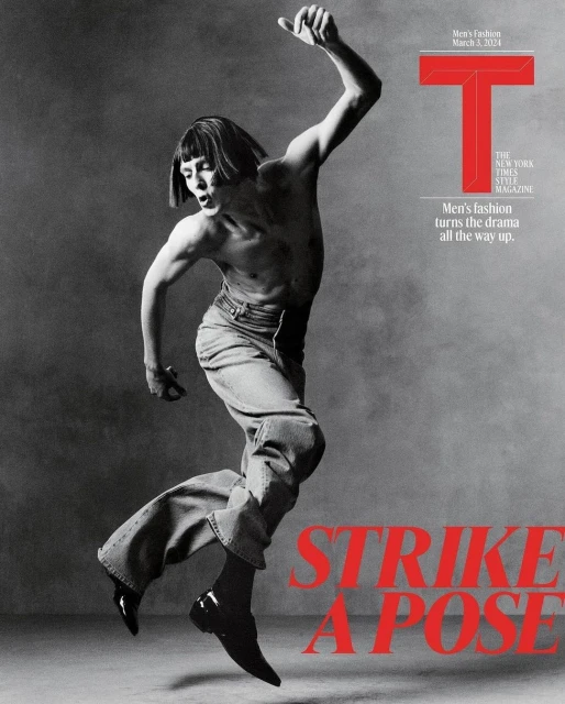 Black and white magazine cover shot of a male fashion model striking a dynamic pose in wide-leg jeans and dress shoes on gray background.