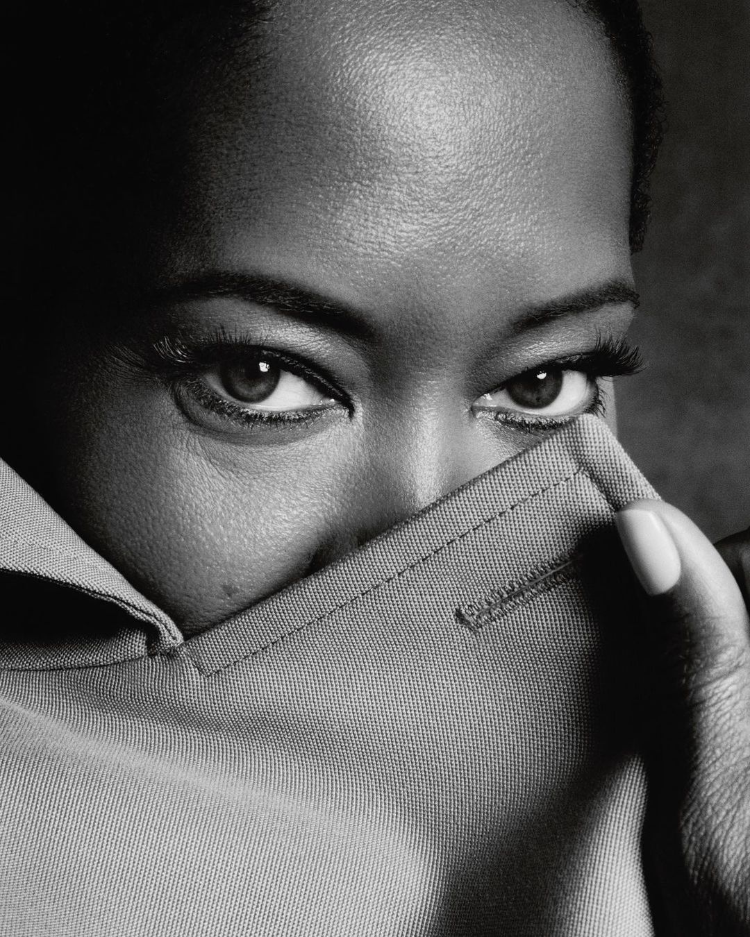 Monochrome close-up of an adult female with dark skin partially covering her face with a light grey collared shirt indoors.