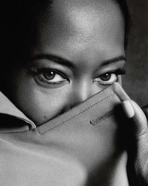 Monochrome close-up of an adult female with dark skin partially covering her face with a light grey collared shirt indoors.