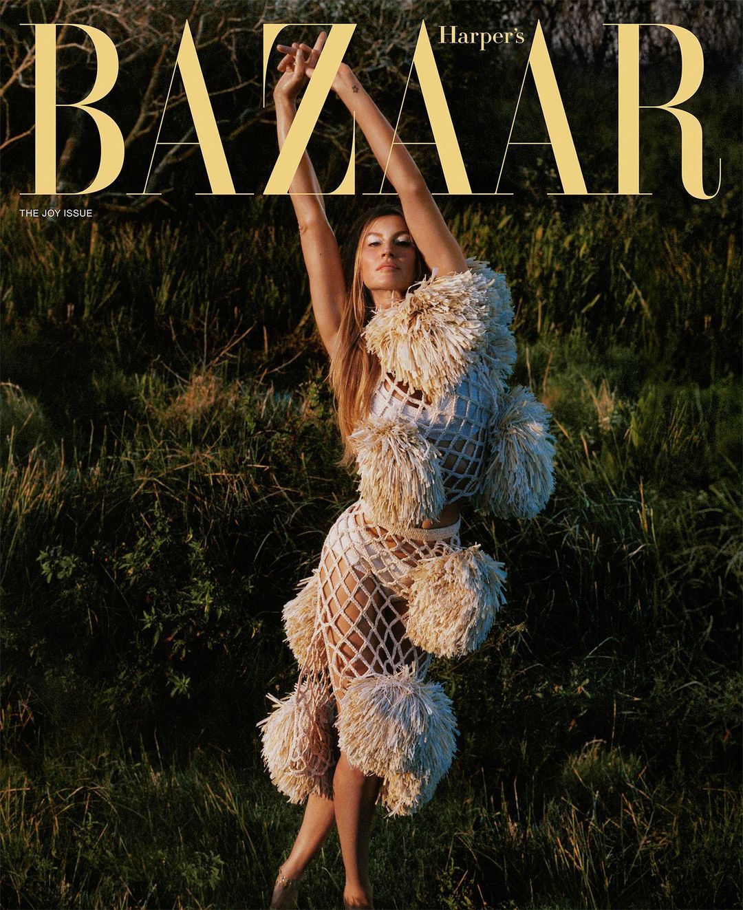 Gisele Bundchen posing outdoors in a textured fringed outfit with arms raised in a grassy field for Harper's Bazaar