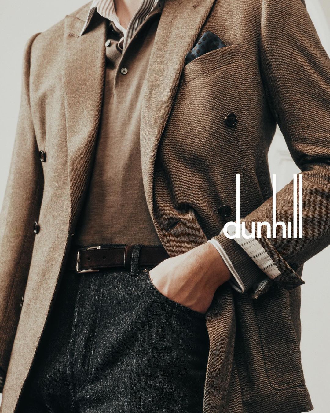 Man in a brown blazer and grey trousers, hand in pocket, with a polo shirt and belt, posing indoors. Dunhill logo is visible.
