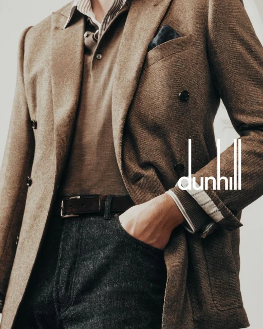 Man in a brown blazer and grey trousers, hand in pocket, with a polo shirt and belt, posing indoors. Dunhill logo is visible.