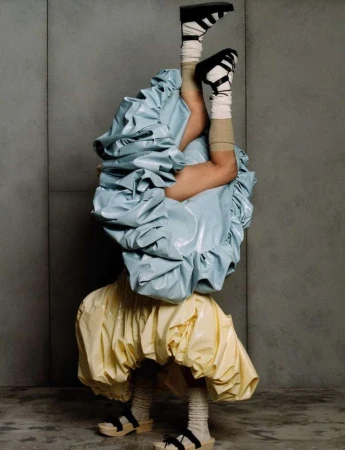 Person in upside-down handstand wearing platform sandals, socks, and two-toned voluminous dresses against a gray wall backdrop.