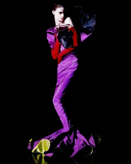 Fashion shot of a woman in a purple gown and red gloves, holding a black purse against a dark background, fashion-forward and bold.