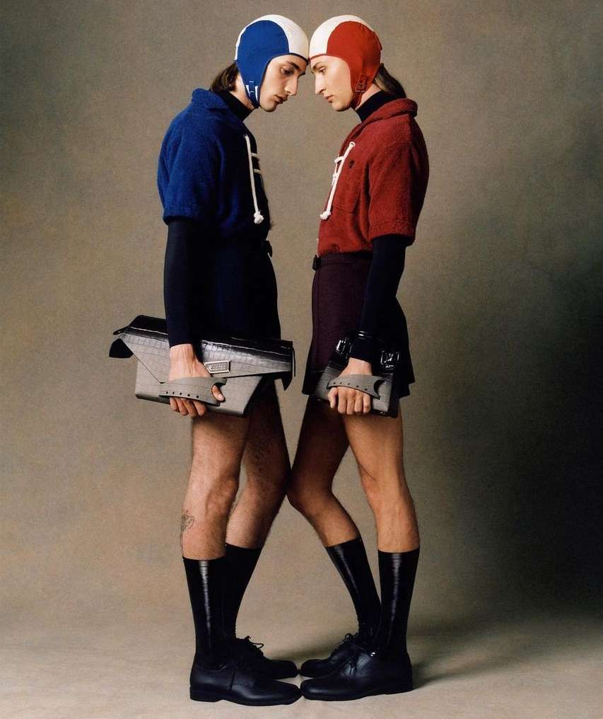 Two fashion models in matching helmets and outfits stand face-to-face holding bags, striking a pose in a studio with a neutral backdrop.
