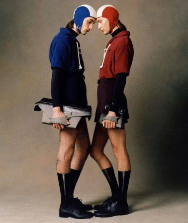 Two fashion models in matching helmets and outfits stand face-to-face holding bags, striking a pose in a studio with a neutral backdrop.