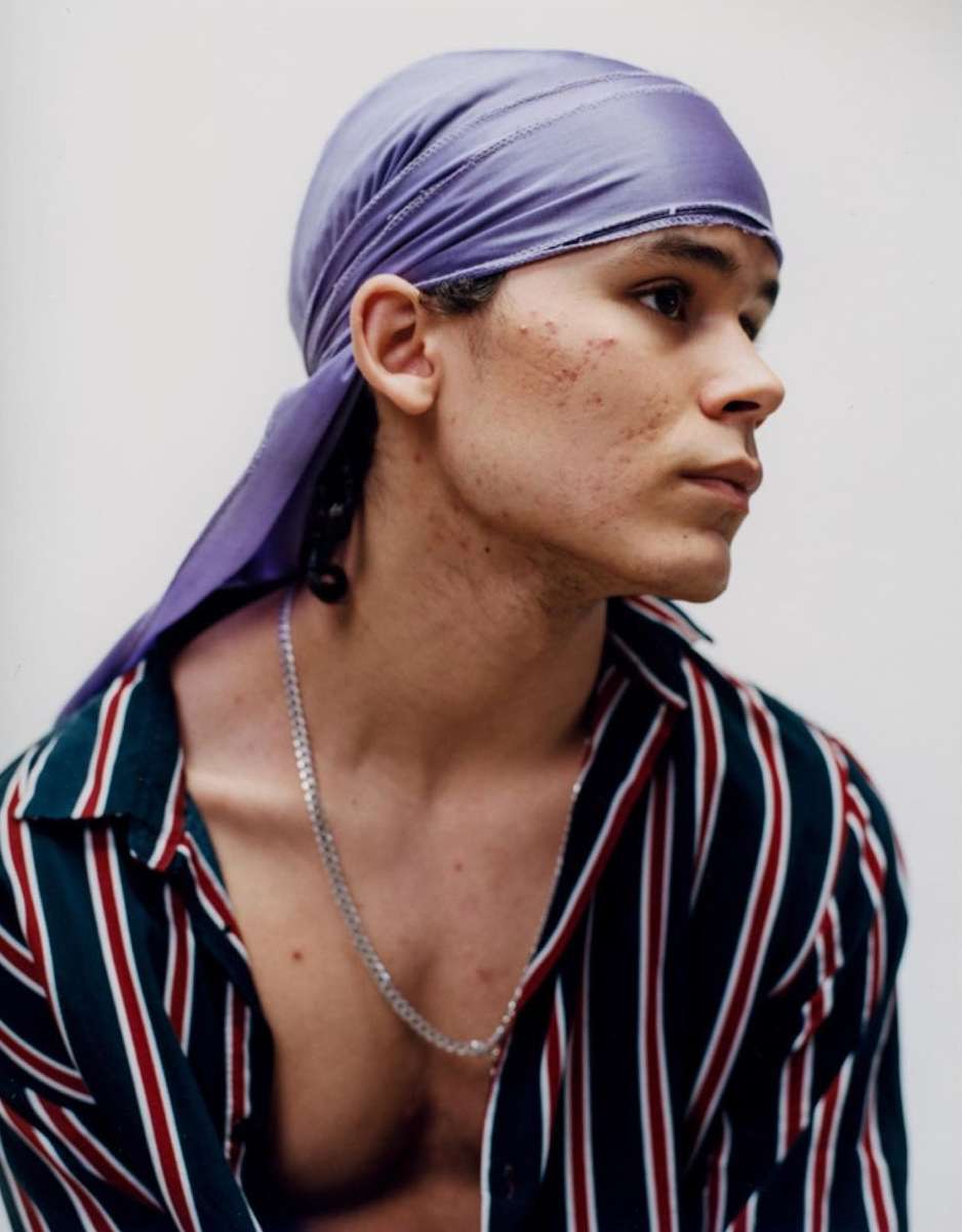 Portrait of a young adult man wearing a purple durag and striped shirt against a light background, showcasing a fashion-forward aesthetic.