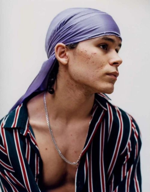Portrait of a young adult man wearing a purple durag and striped shirt against a light background, showcasing a fashion-forward aesthetic.