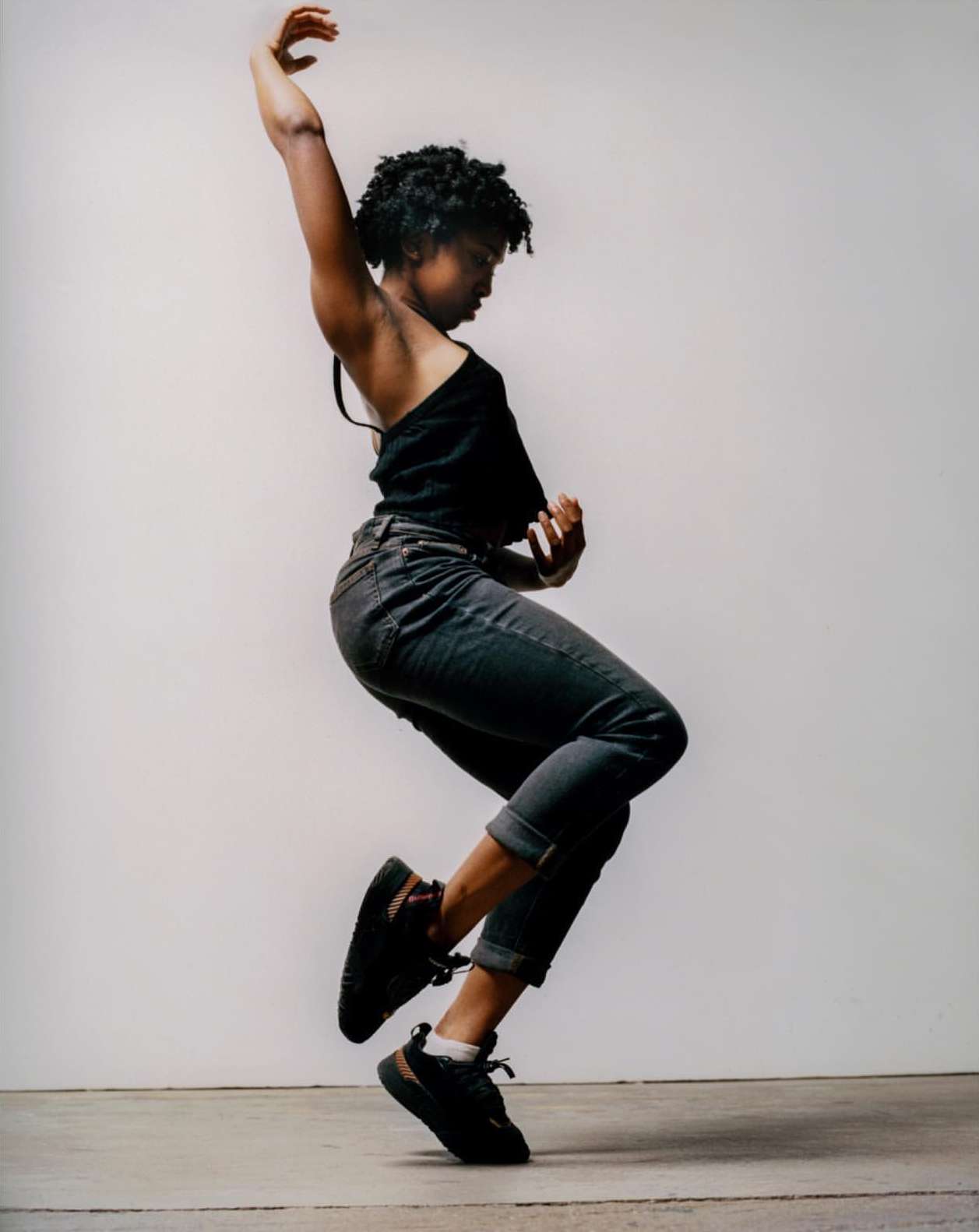 Young woman dances expressively in a photo studio, wearing a black top, rolled-up jeans, and sneakers against a neutral backdrop.