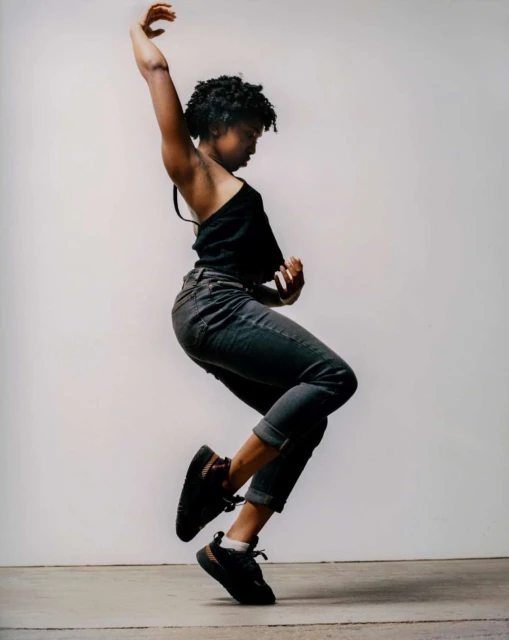 Young woman dances expressively in a photo studio, wearing a black top, rolled-up jeans, and sneakers against a neutral backdrop.