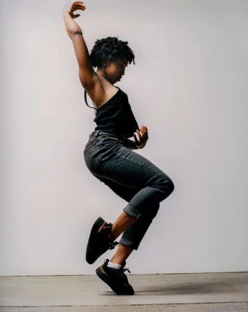 Young woman dances expressively in a photo studio, wearing a black top, rolled-up jeans, and sneakers against a neutral backdrop.