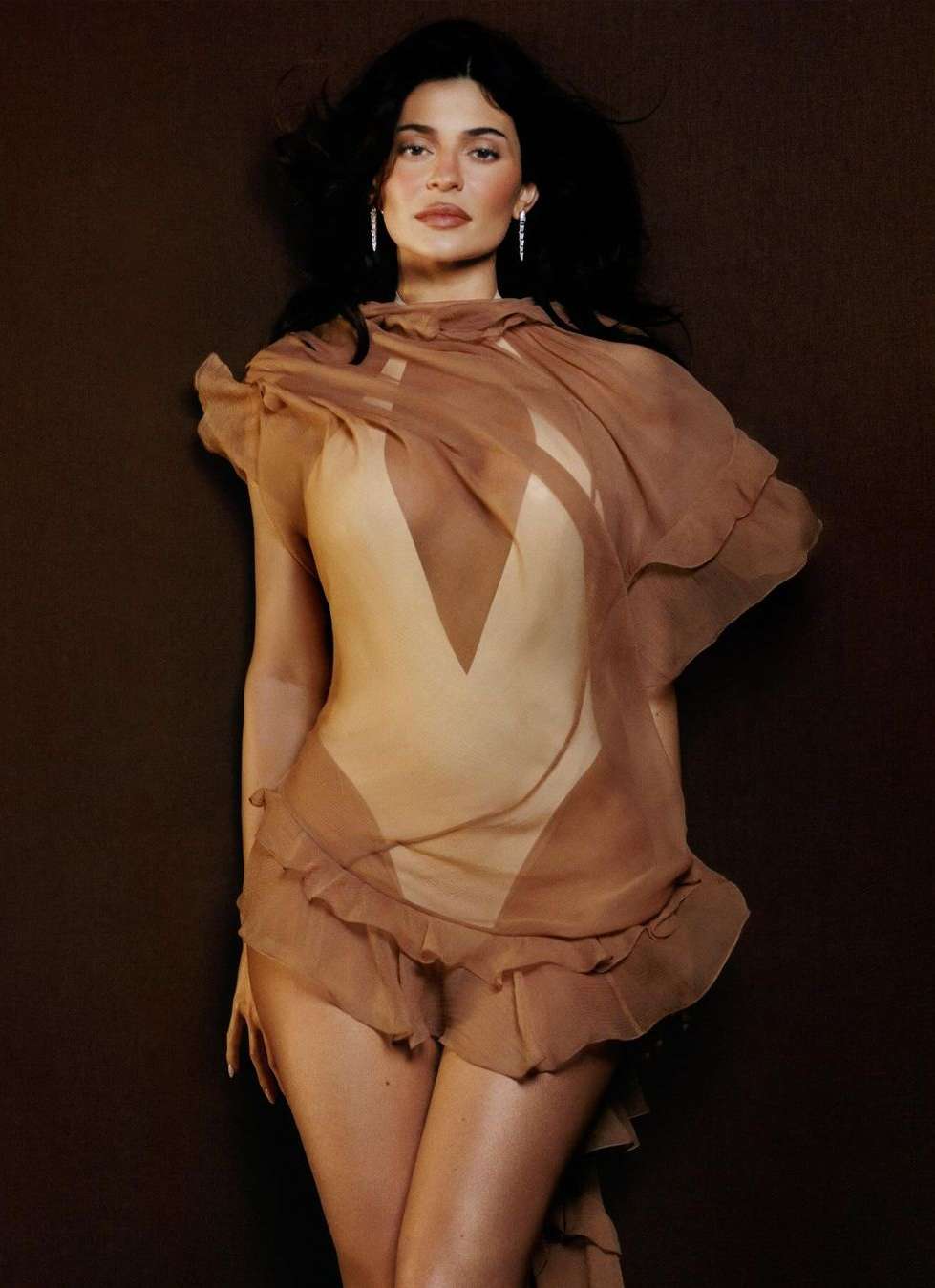 Brunette woman in tan bodysuit with brown sheer fabric detail poses against dark brown backdrop for a fashion or beauty portrait.