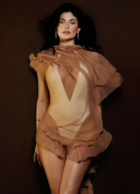 Brunette woman in tan bodysuit with brown sheer fabric detail poses against dark brown backdrop for a fashion or beauty portrait.