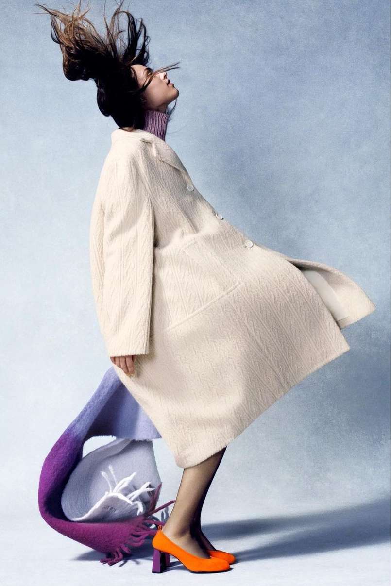 Fashion shot of a person in a cream coat with their hair in the air, wearing a purple turtleneck, orange high heels, and a colorful scarf.