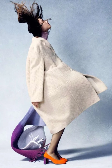 Fashion shot of a person in a cream coat with their hair in the air, wearing a purple turtleneck, orange high heels, and a colorful scarf.