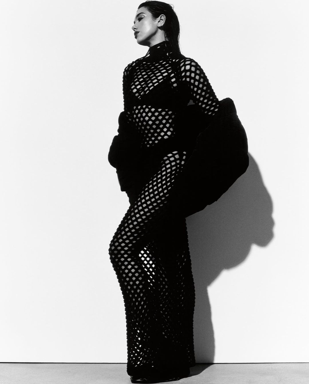 A stylish woman in a black mesh dress poses against a bright white background in a studio portrait. Fashion and elegance meet simplicity.