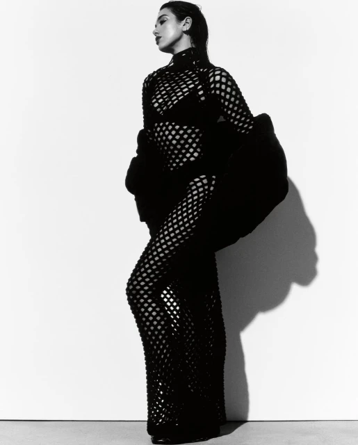 A stylish woman in a black mesh dress poses against a bright white background in a studio portrait. Fashion and elegance meet simplicity.