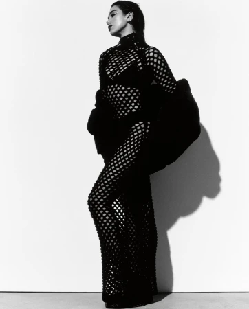 A stylish woman in a black mesh dress poses against a bright white background in a studio portrait. Fashion and elegance meet simplicity.