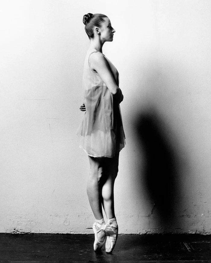 Ballet dancer en pointe in profile wearing a sheer dress against a plain wall