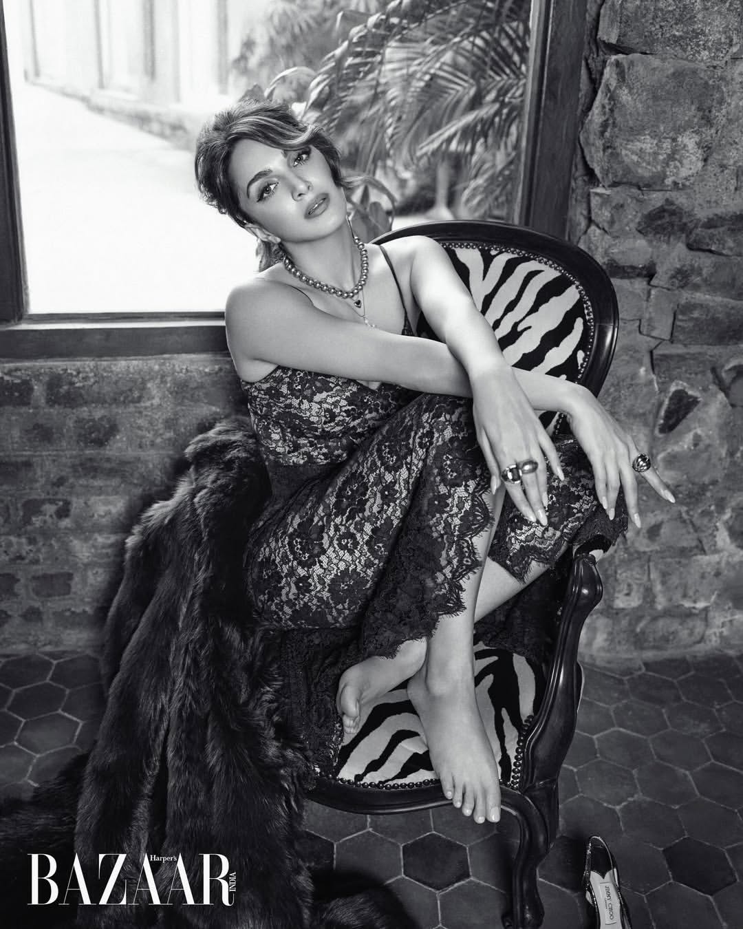 Black and white portrait of an adult woman with dark hair, wearing a lace jumpsuit and jewellery, sitting in a chair with a patterned cushion.