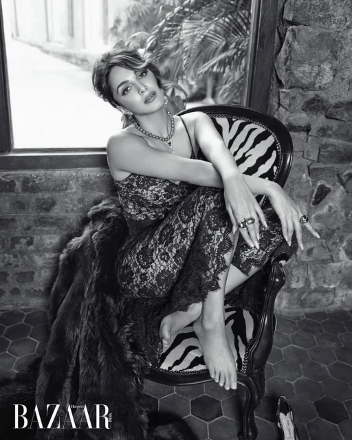 Black and white portrait of an adult woman with dark hair, wearing a lace jumpsuit and jewellery, sitting in a chair with a patterned cushion.
