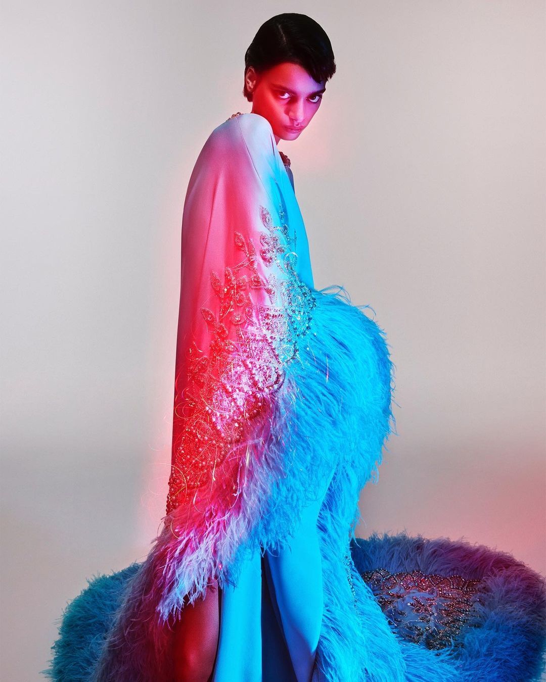 A woman in an elaborate feathered and beaded gown looks directly at the viewer against a light background with red and blue lighting.