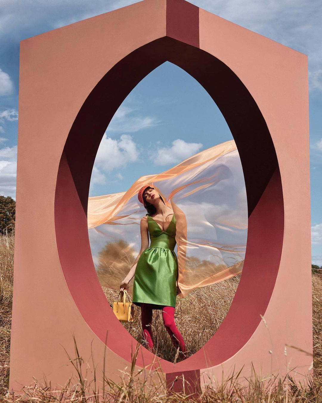 A woman in a green dress and red tights poses within a peach-colored archway in a grassy field, with a flowing peach fabric.
