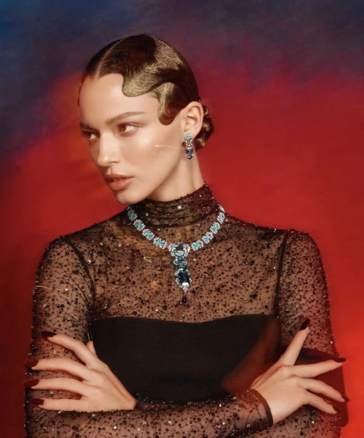 Elegant woman in black gown with blue gemstone jewelry, posing against a red and blue backdrop, showcasing fashion and beauty.