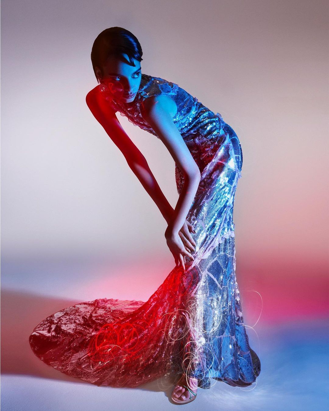 Fashion model poses in a shimmering long gown with blue and red lighting against a neutral backdrop in a studio setting.