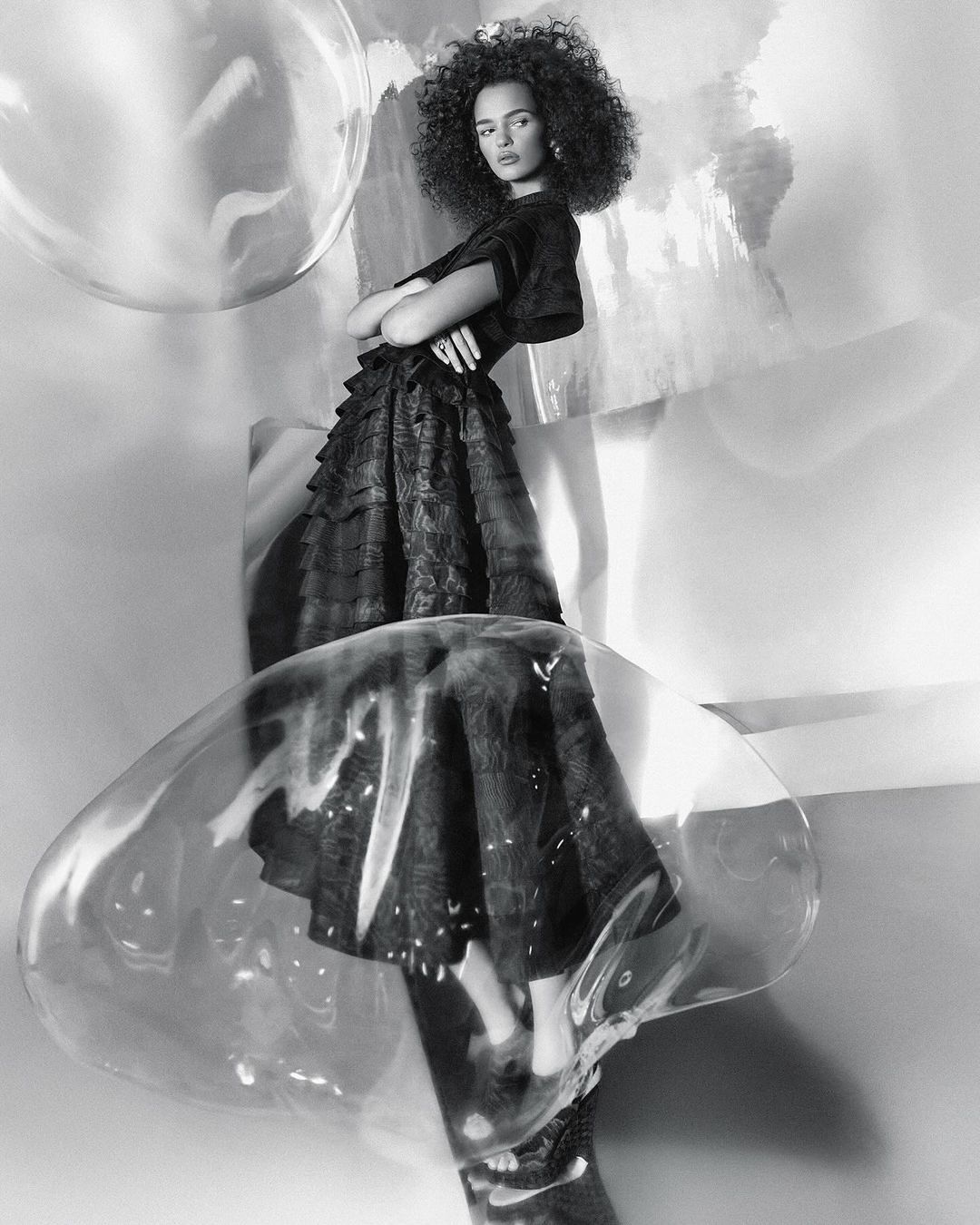 Monochrome studio portrait of a woman with curly hair wearing a dark dress, standing among clear decorative bubbles and abstract shapes.