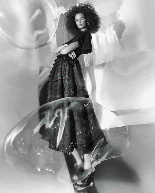 Monochrome studio portrait of a woman with curly hair wearing a dark dress, standing among clear decorative bubbles and abstract shapes.